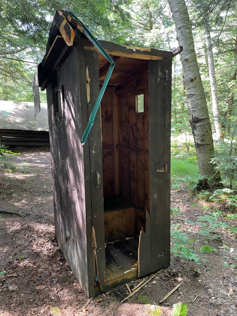 The outhouse behind the lean-to. The door in no slight need of repair.